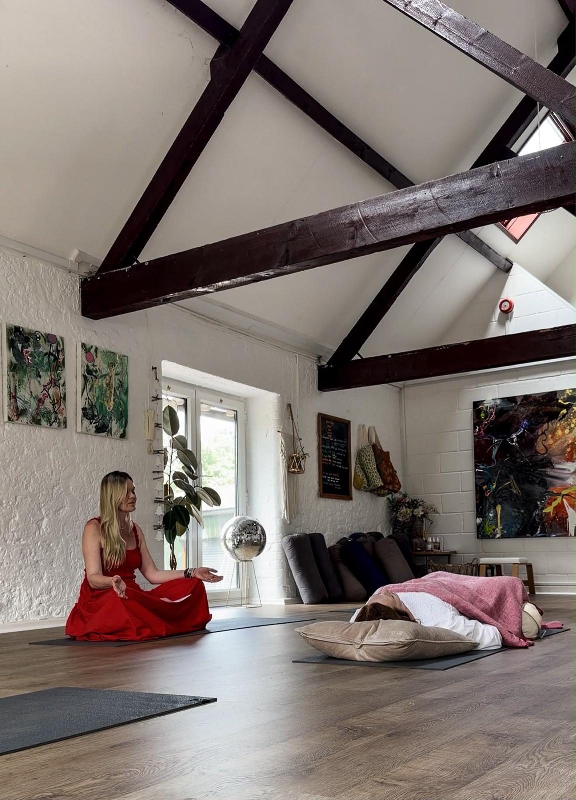 Karina guiding a breathwork session in a beamed studio, with a participant resting under a blanket